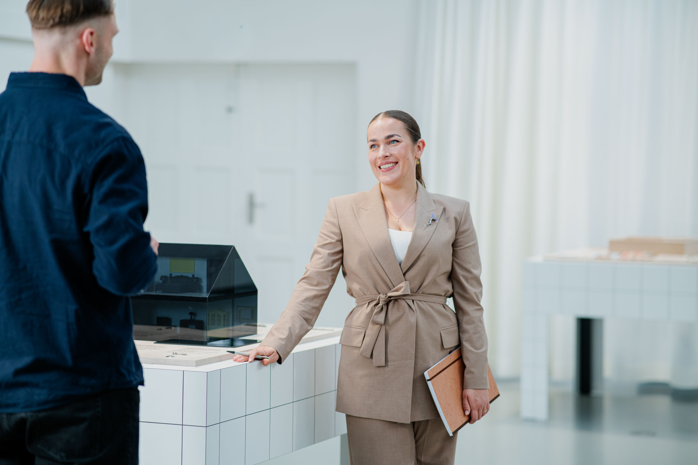 Eine Frau hält ein Klemmbrett und lehnt sich mit der anderen Hand an einen Tisch auf dem ein Architekturmodell steht und lächelt einen Mann an, der vor Ihr steht